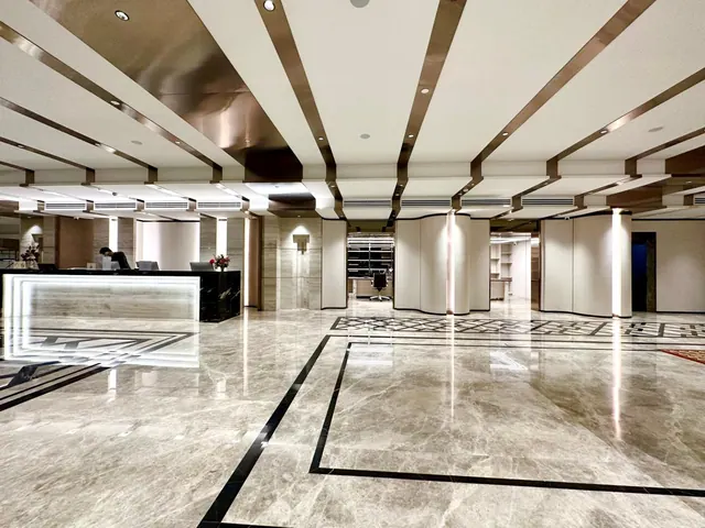 A marble-floored hotel lobby with a illuminated reception desk on the left, a staff member seated behind it, and curved partition walls beyond.