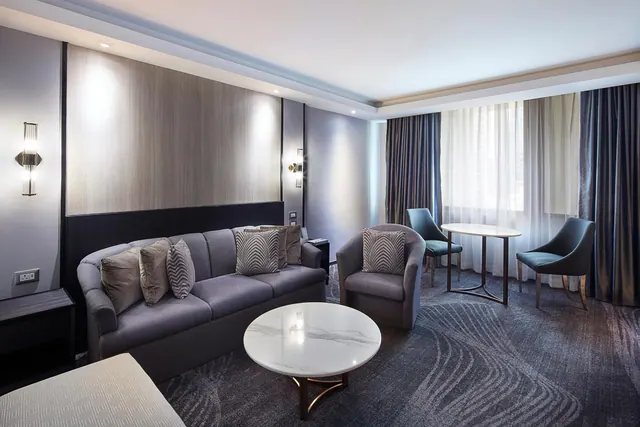 A hotel suite living area with a grey sofa, a round marble coffee table, an armchair, and a small round table with two teal chairs beside a window with sheer curtains.