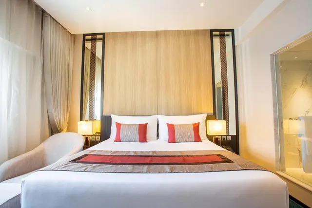 A bedroom with a made king bed, white and red pillows, a wooden headboard, bedside lamps, an armchair, and sheer curtains, with a glass wall showing a marble bathroom on the right.