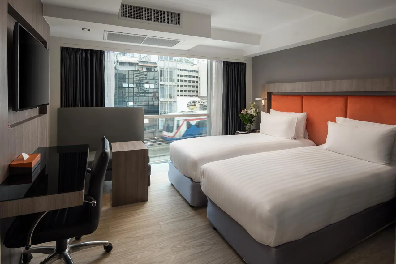 A bedroom with two beds with white bedding and an orange headboard, a desk and chair, a wall-mounted television, and a large window looking onto city buildings and a passing elevated train.