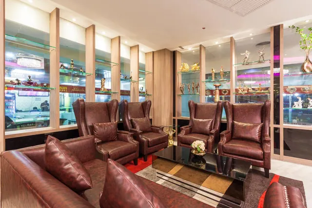 A lounge seating area with brown leather armchairs and a sofa, a glass coffee table with a flower arrangement, and figurines displayed on glass shelves behind.