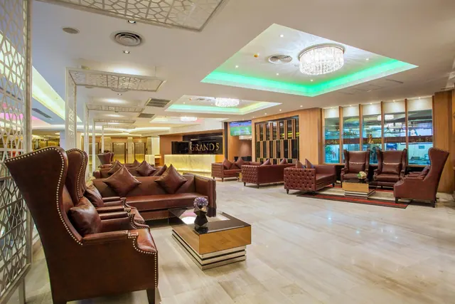 The lobby lounge has leather armchairs and sofas, a reception desk with a GRANDS sign at the back, and a chandelier under a green-lit ceiling.