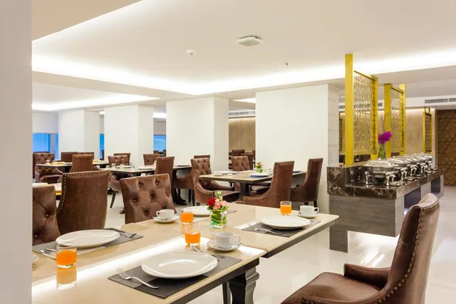 A breakfast dining room with set tables, brown tufted leather chairs, and a buffet counter with chafing dishes on the right.