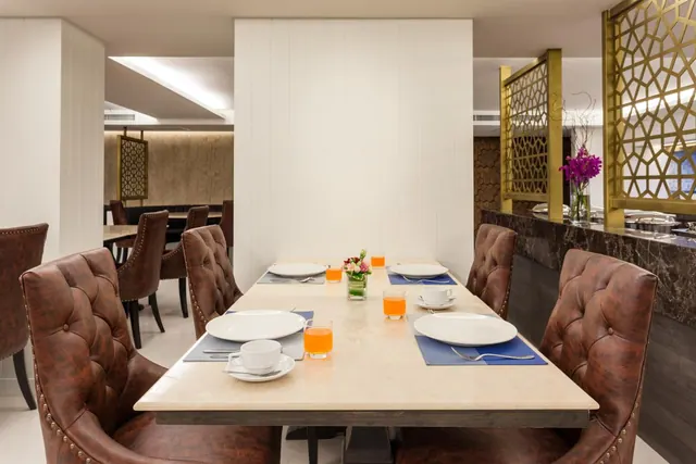 A long marble dining table is set with plates, cups, orange juice glasses and a flower vase, with brown tufted leather chairs and a buffet counter behind gold lattice dividers.