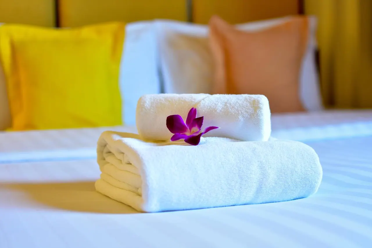 Two folded white towels with a purple orchid on top rest on a bed with yellow, orange and white cushions behind them.