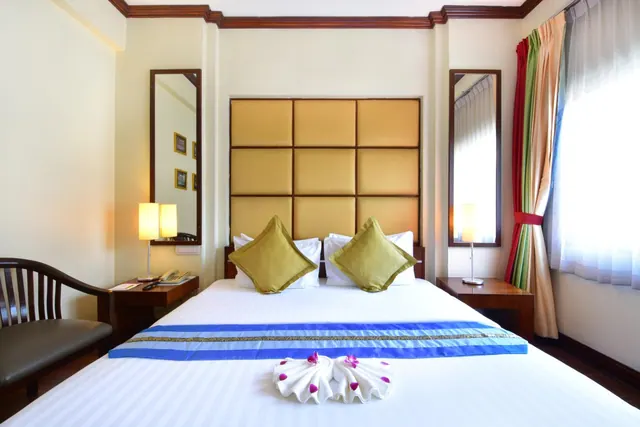 A hotel bedroom with a double bed under a gold padded headboard, olive cushions, a blue and gold bed runner, and towel swans on the duvet, with a wooden chair, bedside lamps and a window with sheer curtains.