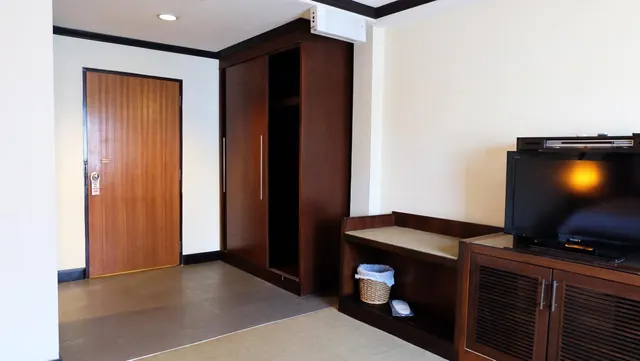 A hotel room entrance with a wooden door, a wardrobe, and a wall-mounted TV on a wooden cabinet beside a shelf holding a basket of towels.