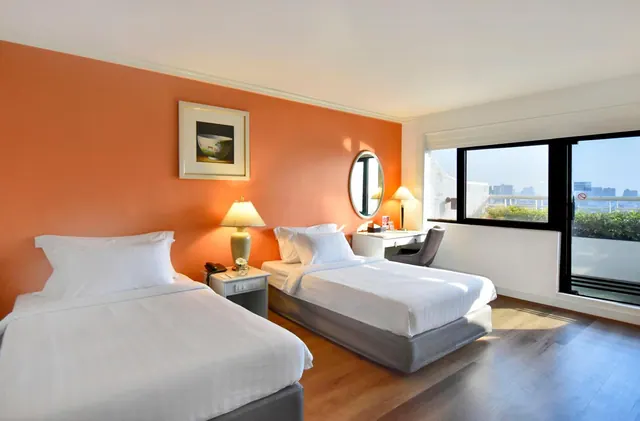 A bedroom with two single beds, an orange accent wall, a desk with a round mirror, and a glass door opening onto a balcony with a city view.