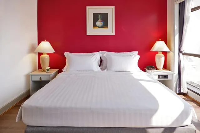 A made-up king bed with white striped linens sits against a red accent wall, flanked by two table lamps, with a curtained window on the right.