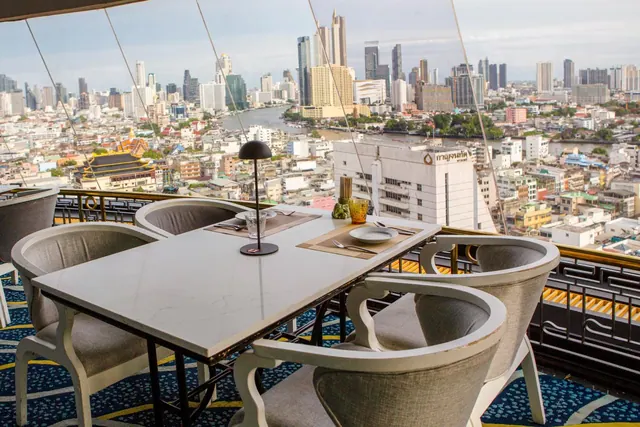 A set dining table with grey upholstered chairs on an open terrace overlooks a dense city skyline and a river.