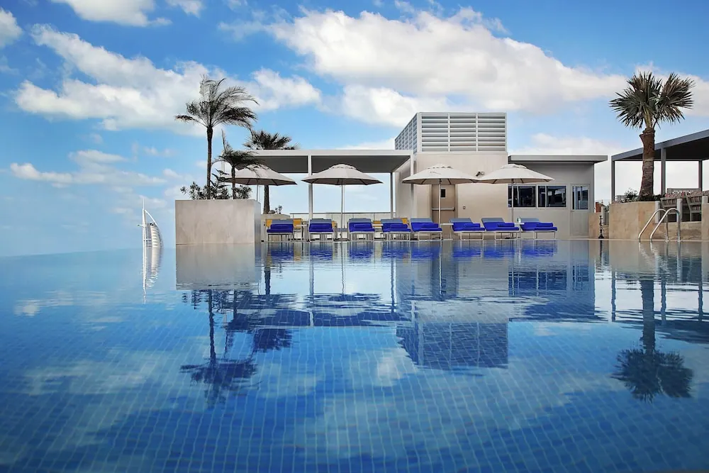 An infinity-edge pool with blue-cushioned loungers and white umbrellas, with the Burj Al Arab visible on the left beyond the water.