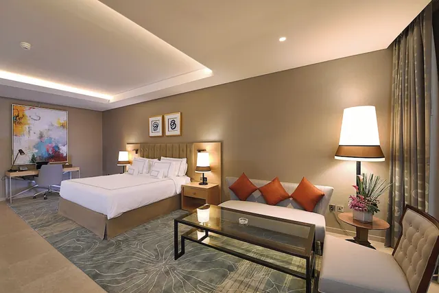 A staged hotel bedroom with a king bed, a seating area with orange cushions, a glass coffee table, a desk and chair, and floor lamps.