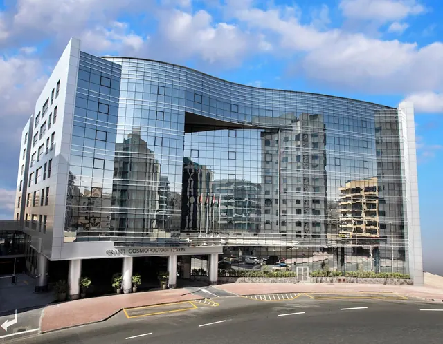 The curved glass facade of the Grand Cosmopolitan Hotel stands under a blue sky, with a covered entrance and road in front.