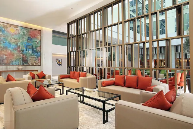 A lobby seating area with beige sofas and armchairs, orange cushions, glass coffee tables, a large abstract painting and a bronze slatted partition.