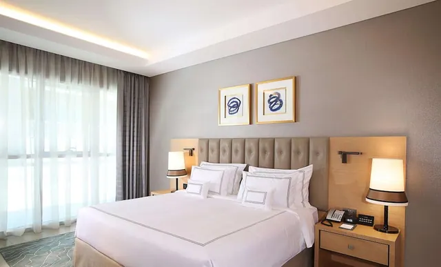 A bedroom with a made-up king bed, tufted headboard, two bedside lamps and sheer curtains over a large window.