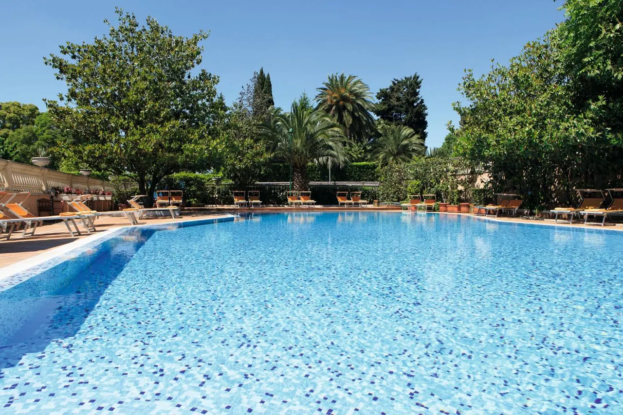 An outdoor swimming pool with blue mosaic tiles is surrounded by sun loungers, palm trees and tall green trees under a clear sky.