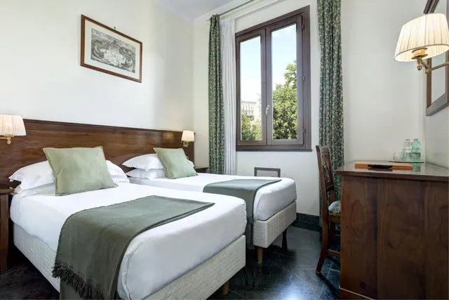 A twin bedroom with two made beds, a framed print on the wall, green curtains by a window showing trees outside, and a wooden desk with bottled water.