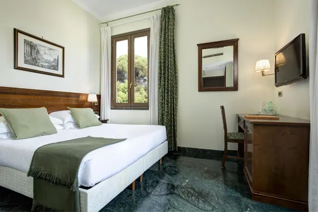 A double bed with green pillows and a green throw sits beside a window, with a wall-mounted television and a wooden desk on the right.