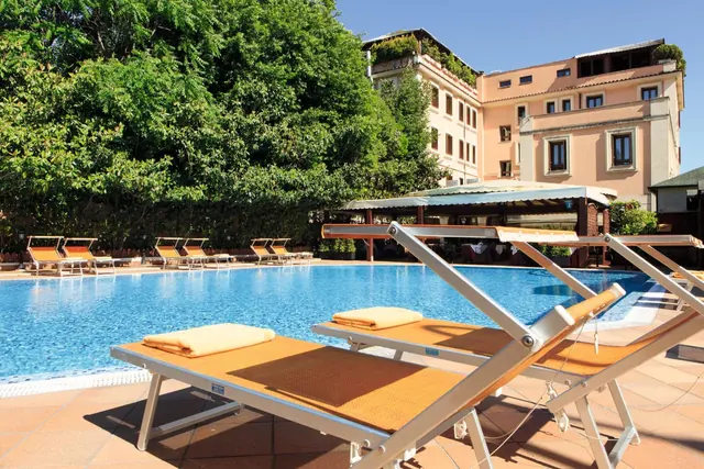 Outdoor swimming pool with orange sun loungers and yellow towels in the foreground, with a pale pink multi-storey building and trees behind.