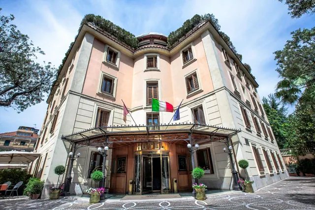 The front of a multi-storey hotel building with a glass entrance canopy, a sign reading HOTEL, and an Italian flag hanging from the facade.