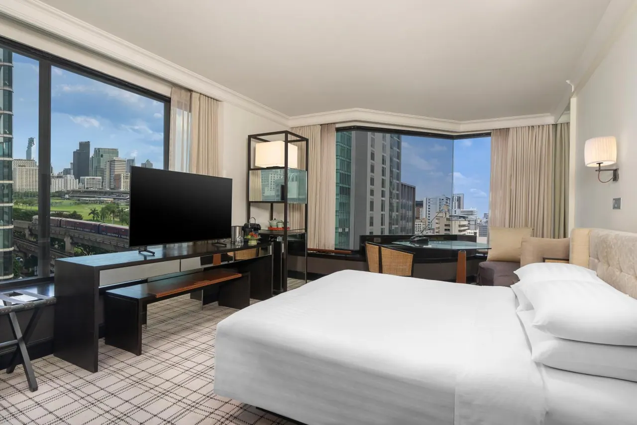 A hotel bedroom with a king bed facing a wall-mounted television, with floor-to-ceiling windows showing a city skyline, a green area and an elevated train line.