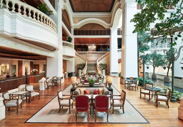 A grand hotel lobby with white colonnades, a curved upper balcony, a central seating area with armchairs and a sofa, and a reception desk on the left.