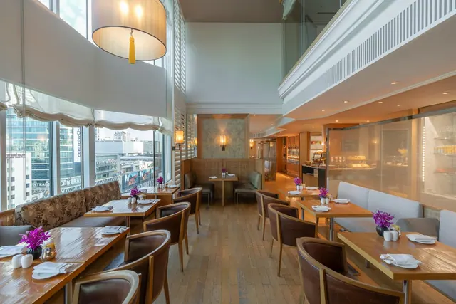 A double-height restaurant dining room with wooden tables, upholstered chairs and booth seating, with large windows showing city buildings outside.