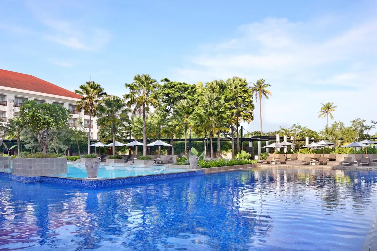 A large blue pool with sun loungers and shade umbrellas along its edge, with palm trees and a red-roofed hotel building behind.