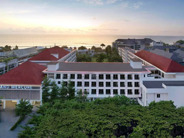 Aerial view of a multi-storey hotel with red-tiled roofs and a partly visible Mercure sign, with the ocean at sunset in the background.