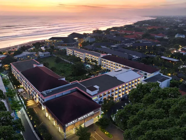 Aerial view at sunset of a multi-storey hotel with illuminated windows beside a sandy beach and ocean, with the Grand Mercure sign at its entrance.