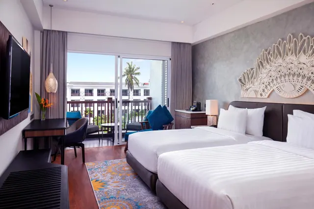 A hotel room with two beds, a desk, a blue armchair and a balcony with sliding glass doors overlooking a white building and palm trees.