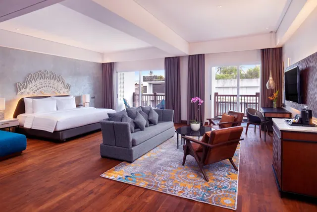 A large hotel room with a king bed, grey sofa, leather armchairs, a desk and a balcony with glass doors onto greenery.