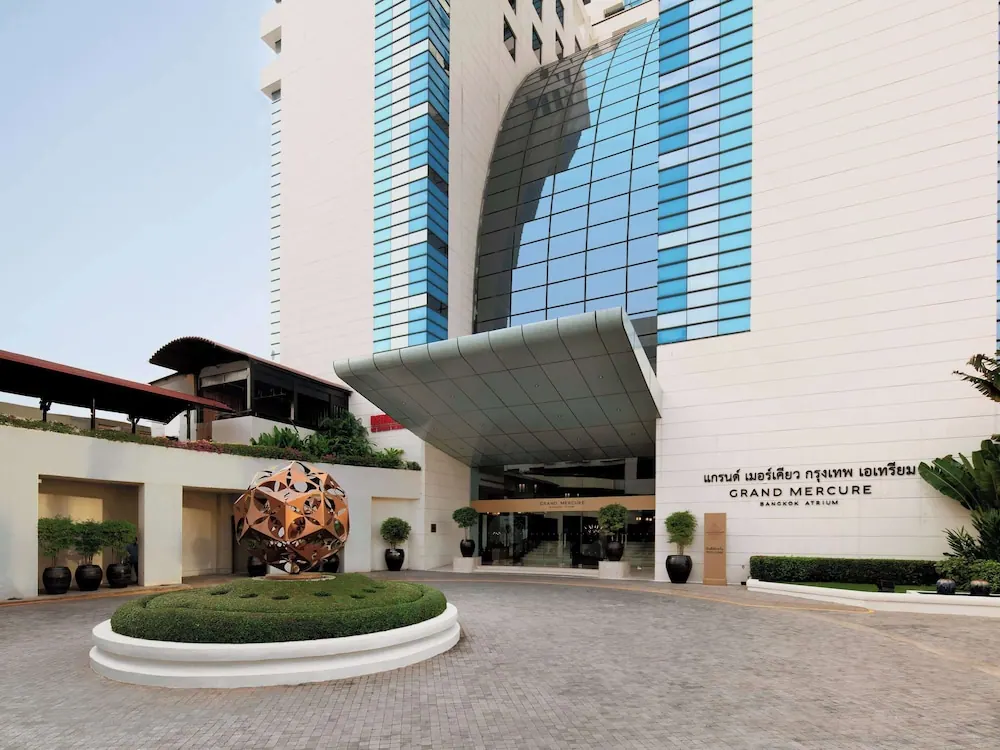 The front entrance of the Grand Mercure Bangkok Atrium hotel, showing a curved glass facade, a covered drop-off canopy, and a circular planted area with a metal sculpture.