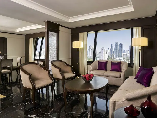 A staged living area with two armchairs, a loveseat and sofa, a round wooden coffee table holding a fruit bowl, and a window showing a city skyline of high-rise towers.