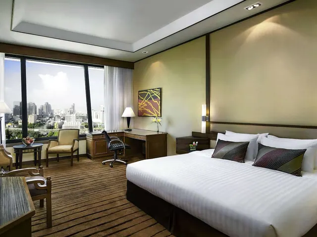 A bedroom with a large white bed, a wooden desk and chair, two armchairs, and floor-to-ceiling windows looking out over city buildings.