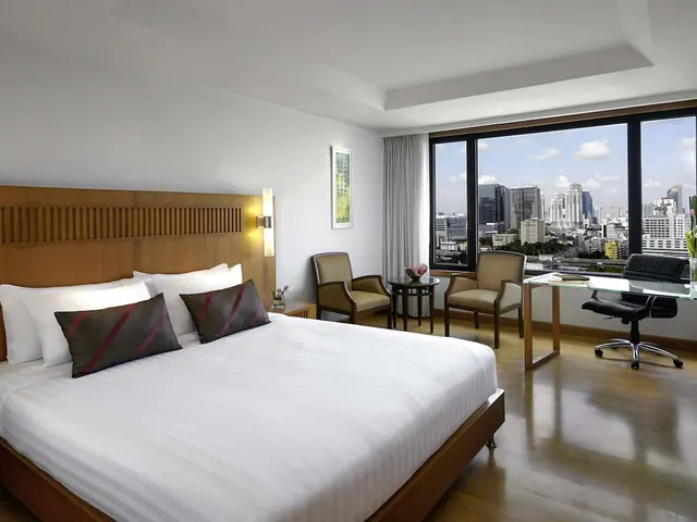 A bedroom with a king bed, two armchairs, a glass desk and a large window looking out over city high-rise buildings.