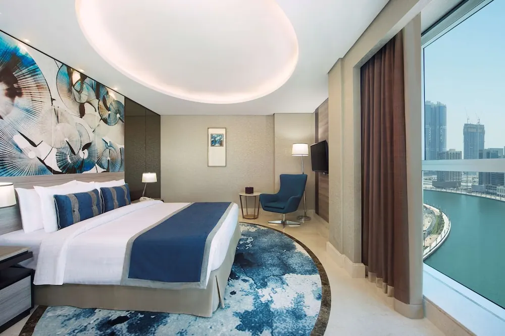 A bedroom with a king bed, a blue armchair, a wall-mounted television and floor-to-ceiling windows looking out over a canal and city towers.