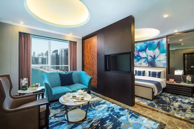 A hotel suite living area with a teal sofa and tea tray on a patterned rug, a bed visible in the background, and a large window overlooking city towers.