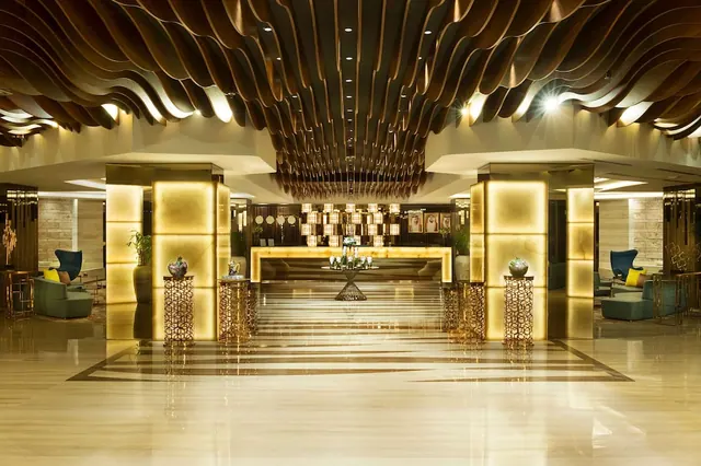 A hotel lobby with a wave-patterned wooden ceiling, illuminated gold columns, a reception desk at the far end and armchairs along the sides.