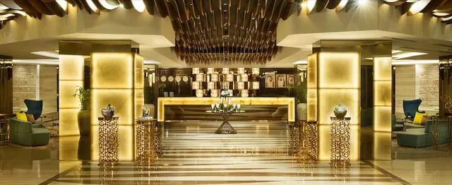 A hotel lobby with illuminated gold-toned pillars, a backlit reception desk, a chandelier cluster and lounge seating on both sides.