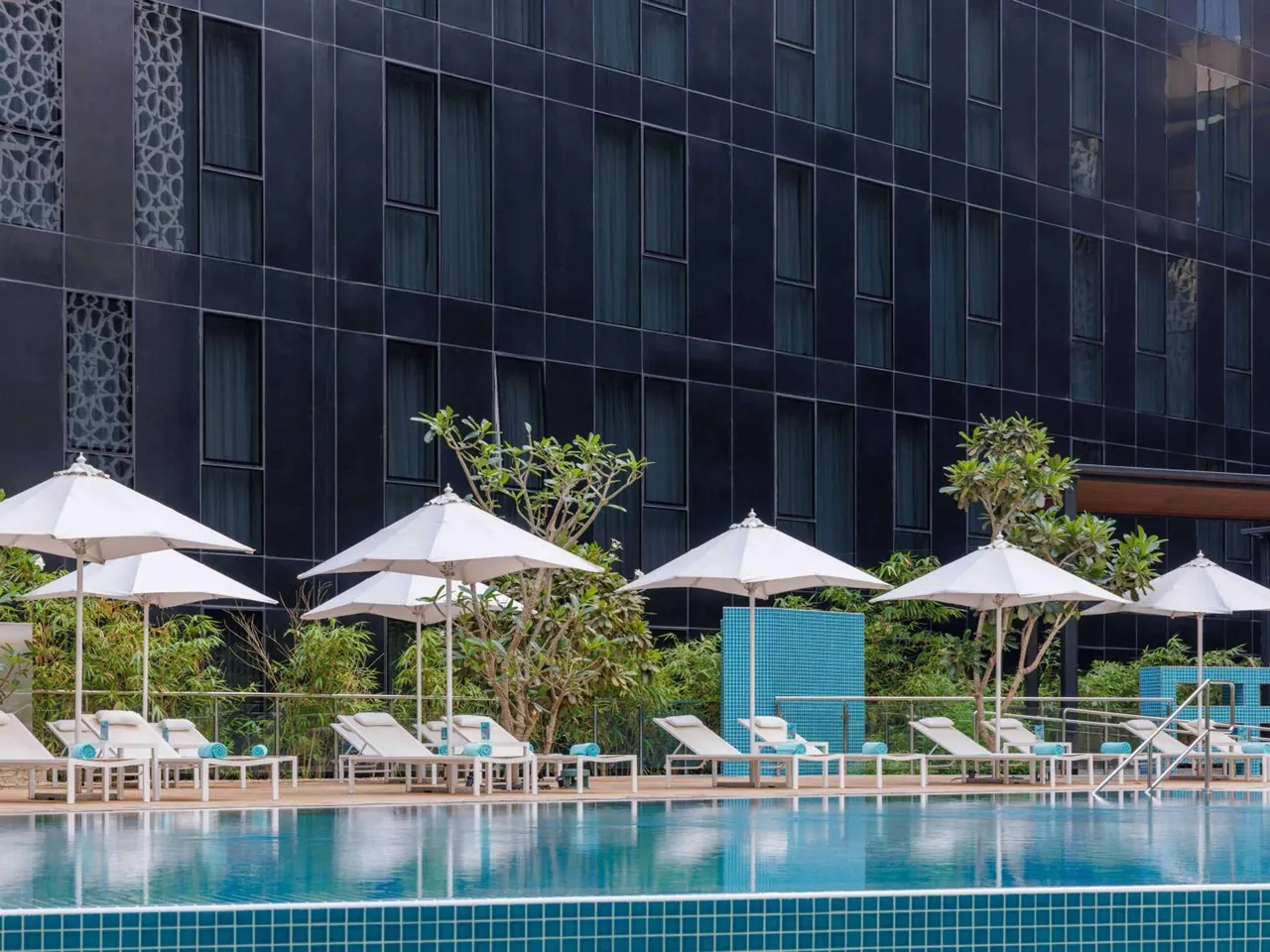 An outdoor swimming pool with white sun loungers, rolled teal towels and white umbrellas on a pool deck, with a dark glass-fronted building behind.