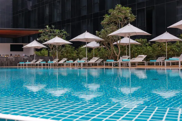 A blue-tiled swimming pool with white parasol umbrellas and sun loungers with teal cushions along its edge, with a dark glass hotel building and trees behind.