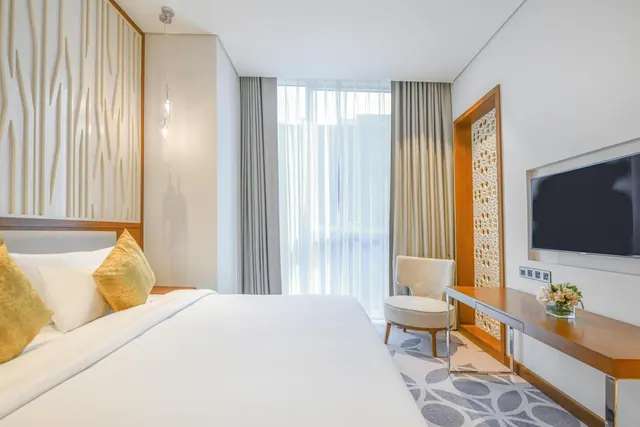 A bedroom with a made white bed with gold cushions, an armchair beside a sheer-curtained floor-to-ceiling window, and a wall-mounted television above a wooden desk.