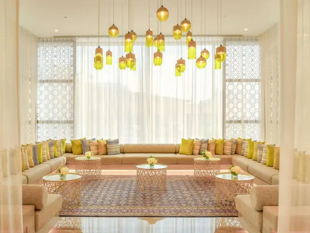 A lounge area with a long curved sofa lined with cushions, round glass-topped side tables with flowers, a patterned rug and yellow pendant lights hanging above.