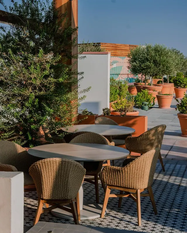 An outdoor terrace with round tables and wicker chairs, surrounded by terracotta planters with olive trees and a wall with a painted mural.
