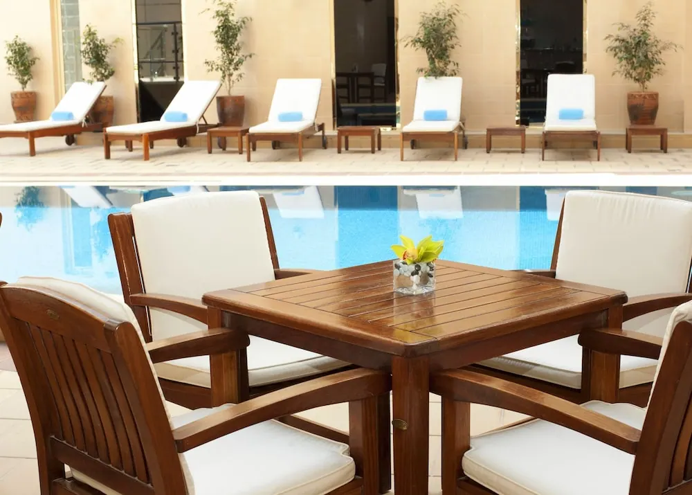 An outdoor pool area with a wooden table and four cushioned chairs in front of the pool, with sun loungers along the far side.