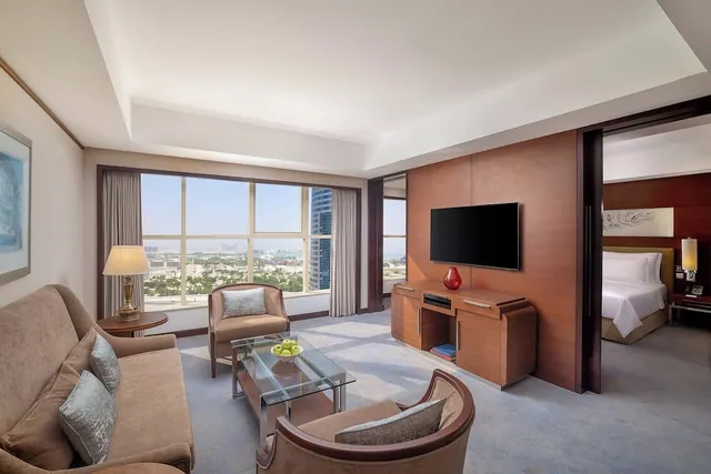 A hotel suite living area with a sofa, armchairs, a glass coffee table, a wall-mounted television and large windows looking over a city, with a bed visible through the doorway on the right.