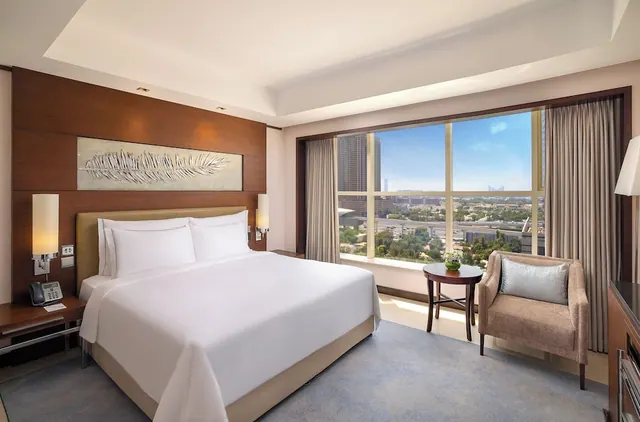 A bedroom with a made king bed, a wood-panelled headboard wall, an armchair and a large window overlooking a city with towers and trees.