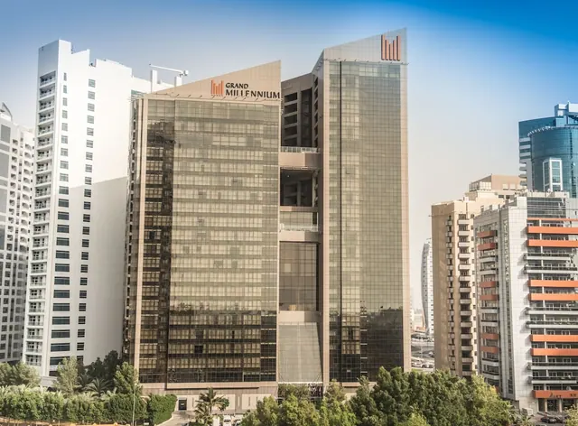 Exterior of the Grand Millennium hotel tower with its glass facade, flanked by residential towers and trees under a clear sky.