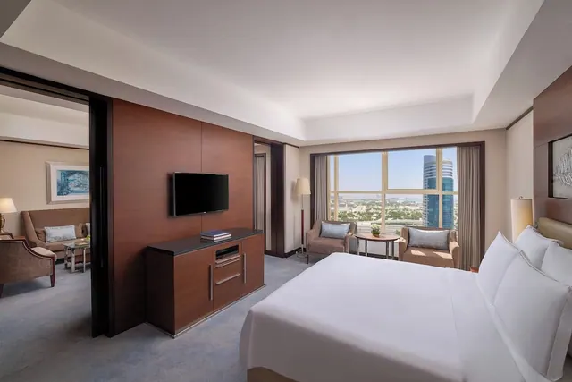 A hotel bedroom with a white king bed, armchairs by a floor-to-ceiling window overlooking the city, and a wall-mounted television above a wooden cabinet.